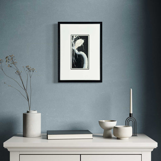 Egret on a Snow-Covered Branch, after Koson. Fine art print of a white egret perched on a snowy branch. Mounted and framed in black, ready to hang. In room set with blue wall.