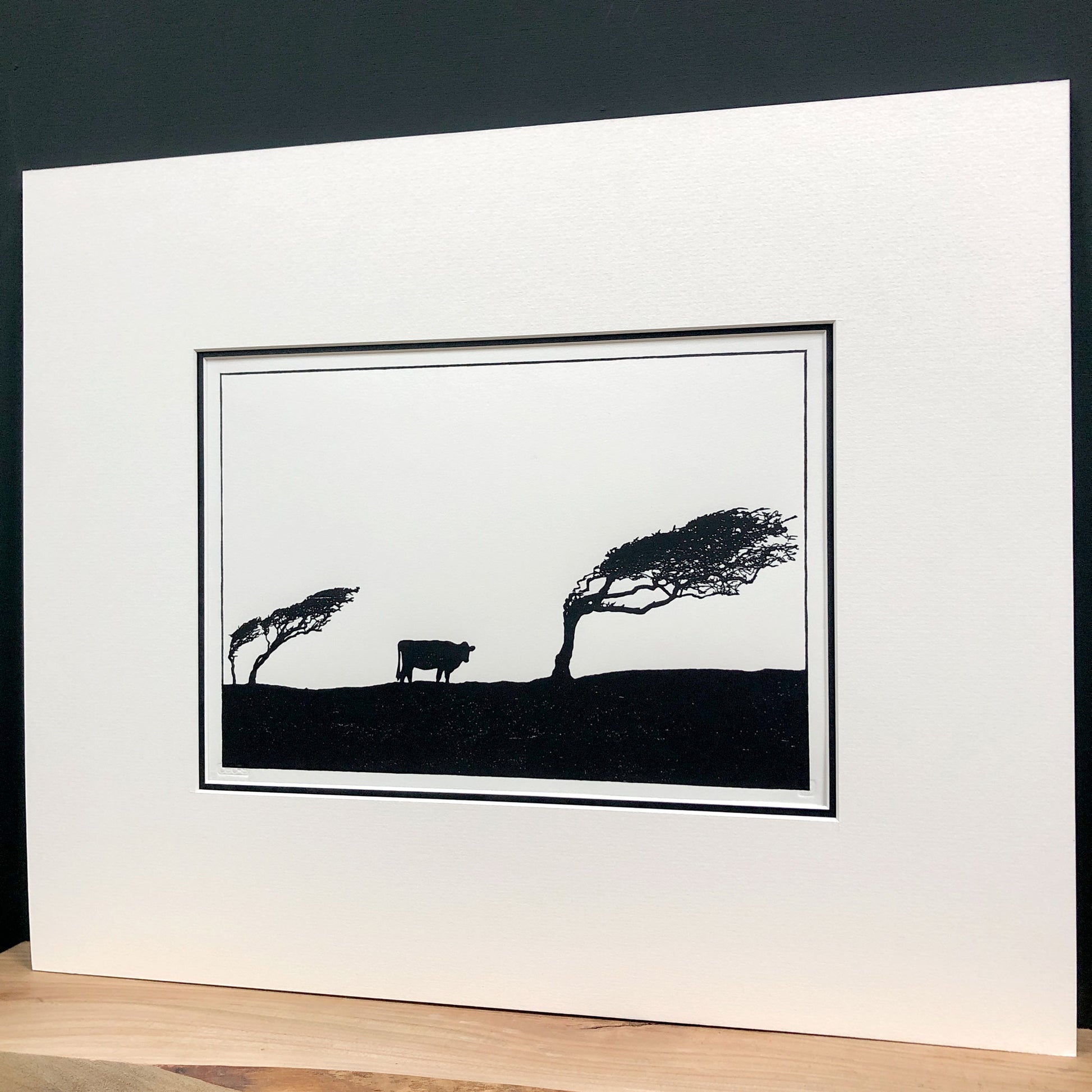 Angled view of mounted windswept hawthorn tree woodblock print showing depth of mount and minimalist black silhouette landscape.