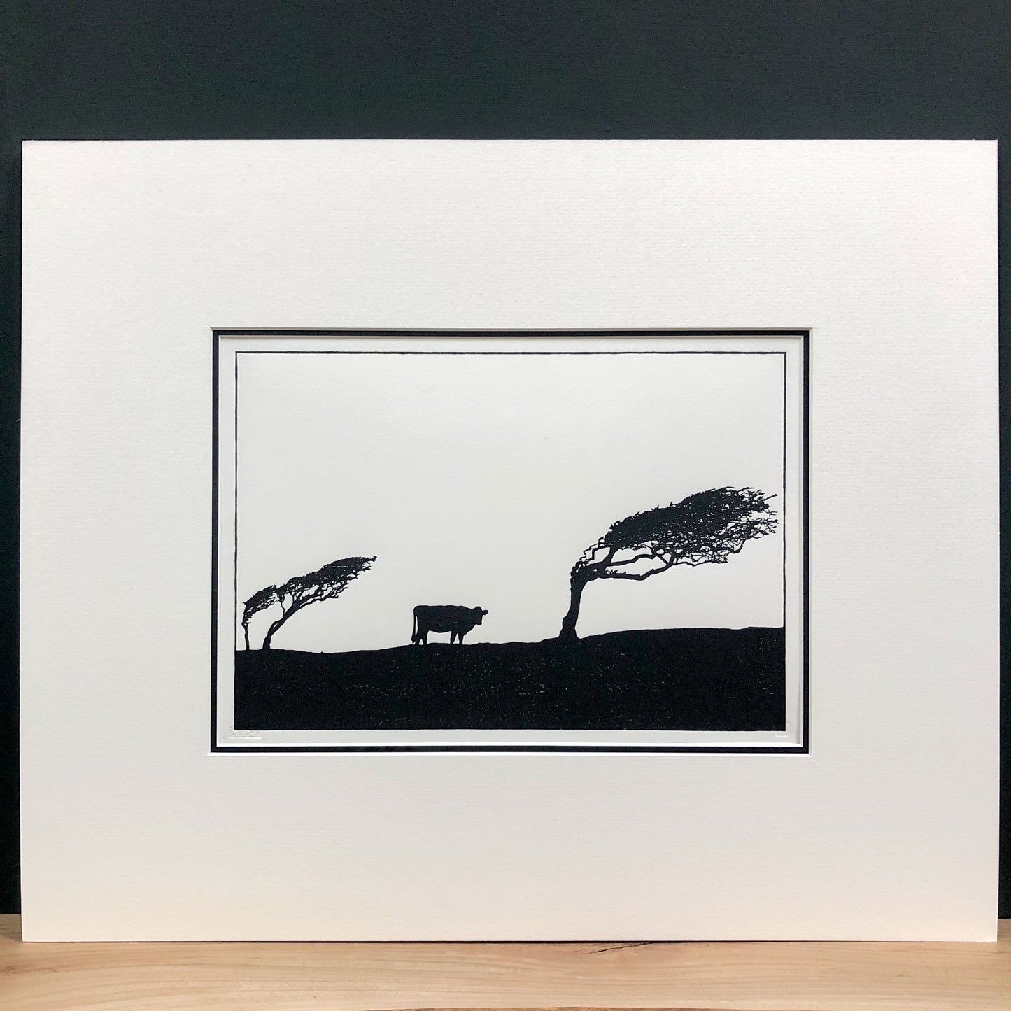 Original handmade woodblock tree print of windswept hawthorns and cow in black ink on cream paper, fourth print in a series of four, mounted with wide cream mount and black inner mount.